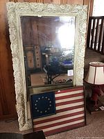 Full view of the large filigree wood framed mirror standing against a wood-paneled wall, with the rustic wooden flag art in front of it.