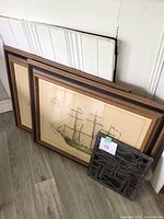 Photo showing two wood framed ship prints stacked against a wall along with a smaller stained glass piece in the front.