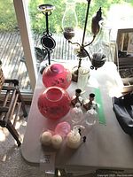 Top view of all lot items on a white table near window showing pink lamp globes, glass vase, candle holders, bottles and figurines.