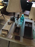Photo showing all items on table including metal base lamp with fabric shade, plaster wall sconce, framed cherub medallion, wood boxes, blue glass bottle, bell, and glass candle holders.