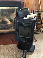 Photo of metal oven with glass door, cast iron kettle on top, and fire starters placed beside it. Oven shows latch and vent panel, black finish.