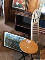 Photo showing white wicker framed mirror, wooden bookcase, folding wood stool, framed painting, needlepoint art, and upholstered stool in corner of room.