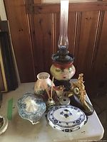 Overview of the lot showing oil lamp, porcelain dishes, vases, clock, candlestick, and figurines on a table.