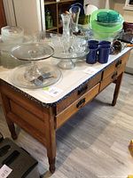 Angle view of rectangular wooden table with black and white speckled metal top, two drawers visible under the top surface, some glassware and dishes on top not included.