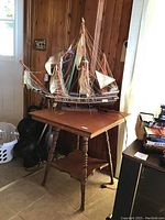 Wooden table with turned legs and triangular base shelf, used condition with scratches. Model ship on table with sails and rigging visible, some unattached strings.