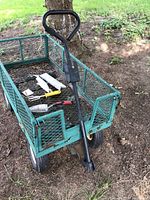 Wide angle photo of green metal garden wagon loaded with small gardening tools and wood sign, placed outdoors on soil by a tree.