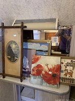 Five framed wall art pieces arranged on a table against a textured wall, showing overall lot content and style variety.