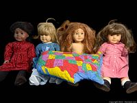 Four American Girl dolls sitting side by side wearing various outfits with a colorful fabric map of the United States laid across their laps.