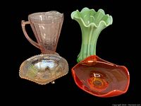 Full lot overview showing five colored glass items against a black background: pink pitcher, tall green ruffled vase, amber carnival glass bowl, pink art glass bowl, and green pressed glass bowl; all items clearly visible