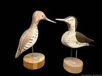 Two hand-carved and hand-painted shorebird decoys standing on round wooden bases, one light brown and one white with brown markings, against a black background.