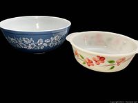 Two vintage mixing bowls, blue Pyrex bowl with white floral pattern and white Fire-King bowl with floral design, displayed on black background.
