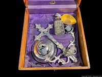 Opened wooden box with purple fabric lining containing various sacrament items including a silver cross, candle holder, etched glass container, and brass bowl.