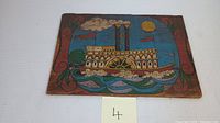 Front perspective of the rectangular wooden plaque featuring a colorful folk art painting of a paddle steamboat with smokestacks, a bright yellow sun, and floral ornamental border.
