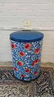 Front-facing photo of a metal kitchen trash can with a blue lid and floral body.