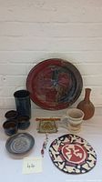 Full view of the studio pottery collection showing all items including platters, cups, vase, mug, pin, and trinket tray on white surface against white brick wall.