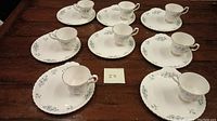 Eight sets of white porcelain cups and saucers with light blue floral patterns and gold trim arranged on a wooden table.