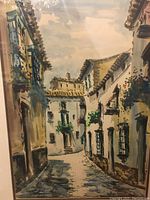 Photo of watercolor painting showing a narrow European street with white buildings, green shutters, and flowers.