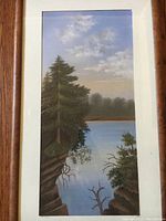 Framed landscape painting depicting trees and water with a cloudy sky. Shows the entire frame and mat with a vertical orientation.