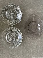 Three Early American Pressed Glass (EAPG) plates laid out on a grey surface. Plates show embossed designs including a crown with Diamond Jubilee text, a coat of arms surrounded by colonial names, and a plate with circular patterns. One plate has edge damage.