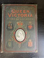 View of the front cover of the antique book showing embossed title text, heraldic shields, and central oval portrait of Queen Victoria.
