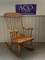 Full view of antique wooden rocking chair with spindle back, curved arms, and gently curved rockers. Chair sits on wooden floor against paneled wall with ACO Antiques & Artifacts sign in background.