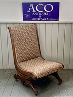 Full view of the rocking chair showing its wooden frame and patterned upholstery against a wood paneled wall with ACO Antiques & Artifacts sign