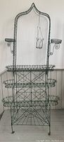 Full frontal view of green painted wire rack showing three tiers, scrollwork sides and hanging planter