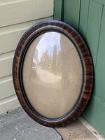 Wooden oval frame leaning against green door and beige siding, showing convex glass and frame finish