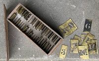 Wooden box open showing brass interlocking stencils organized in slots, some loose stencil plates beside the box