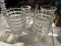 Four clear glass urns or vases with textured geometric patterns and square pedestal bases arranged on a table surface shown under natural lighting.