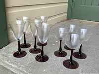 Eight glasses including four wine glasses and four martini glasses with purple stems and bases, clear swirled bowls, arranged on the ground outside near walls.