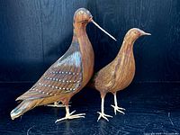 Photo showing the pair of bird figures from a frontal angle highlighting the longer beak of one bird and painted feather details.