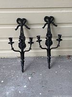 Pair of decorative black iron wall candle sconces with bow and grapevine design, each having three candle holders, displayed against a wall.