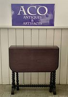 Closed small antique folding wooden table with spool-turned legs under an ACO Antiques & Artifacts sign.