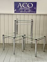 Three vintage mid-century modern chrome and glass side/accent tables positioned together on a wooden floor with white shiplap wall background and ACO Antiques & Artifacts sign above.