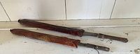Two vintage African ceremonial tools with metal blades and wooden handles inside reddish-brown leather sheaths, laid on white surface.