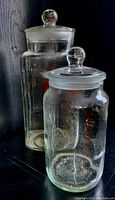 Two clear vintage glass apothecary jars with lids, standing side by side showing overall condition and size difference.