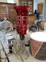 Photo showing vintage cranberry glass lamp with geometric painted decoration on the shade and clear glass pedestal base, with surrounding items for context.