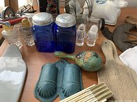 View of the vintage and antique kitchen and household items including glass salt and pepper shakers, ceramic cow creamer, wooden darning egg, linen towel, vintage utensils, and metal kitchen tools.