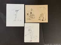 Three black ink cartoon drawings on paper, placed on black surface, showing different characters and signed by George Feyer on one.