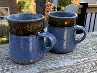 Pair of handmade vintage pottery mugs with speckled blue glaze and dark brown rims on wooden surface outdoors.