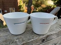 Pair of white ceramic coffee mugs on outdoor wooden surface, showing blue 'portobello road' and red 'notting hill' text markings on the sides.