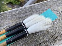 Close-up image showing three horsehair brushes with white bristles, black lacquered handle sections, and original blue card still attached.