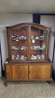 Front view of antique wooden display cabinet with glass doors, clawed feet, and multiple shelves inside containing china cups and plates (not included in lot).