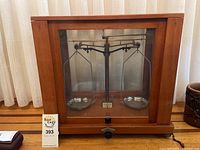 Front view of entire balance scale inside wooden display case with glass panels showing metal pans and scale mechanism.