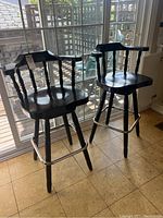 Pair of black wooden swivel bar stools with arm rests and metal footrests photographed indoors near a sliding glass door with patio outside visible.