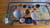 Glass top table displaying various brass, pewter, glass, and ceramic dishes and decorative items arranged neatly for viewing.