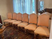 Six upholstered dining chairs lined up against a window with sheer curtains, illustrating overall condition, fabric color, and wooden legs.