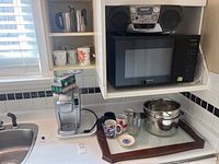 Wide view showing Keurig coffee maker next to several mugs on a serving tray, and a pot with strainer on countertop under microwave.