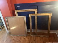 Three large wooden frames, two with glass, arranged on a hardwood floor against walls